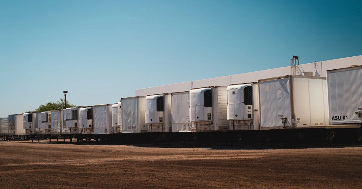 A row of white semi-truck trailers parked on dirt ground under a clear blue sky. 