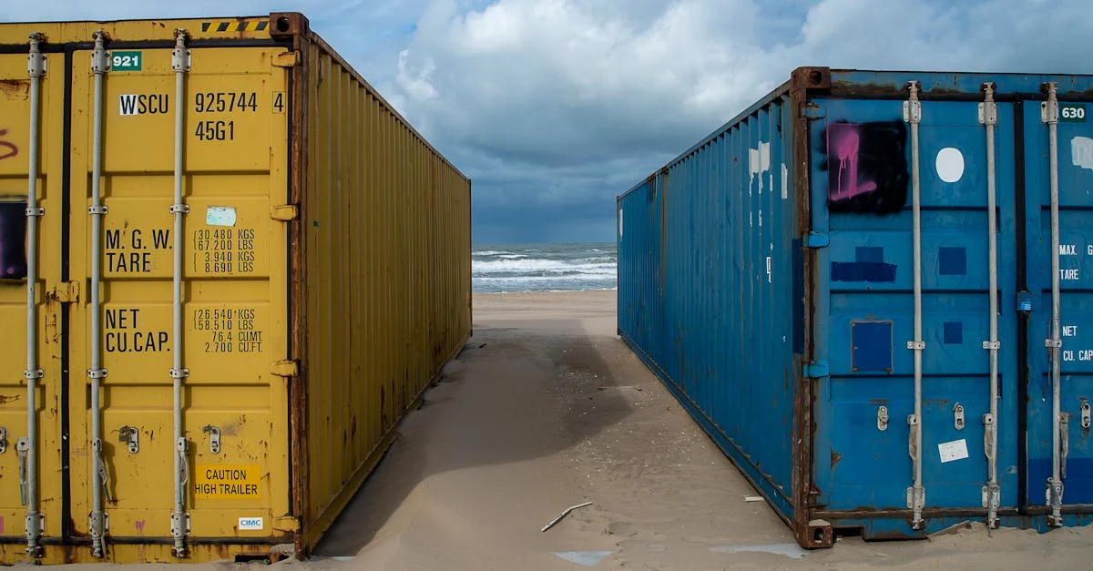 Two large shipping containers, one yellow and one blue, stand on a sandy beach under a cloudy sky. 