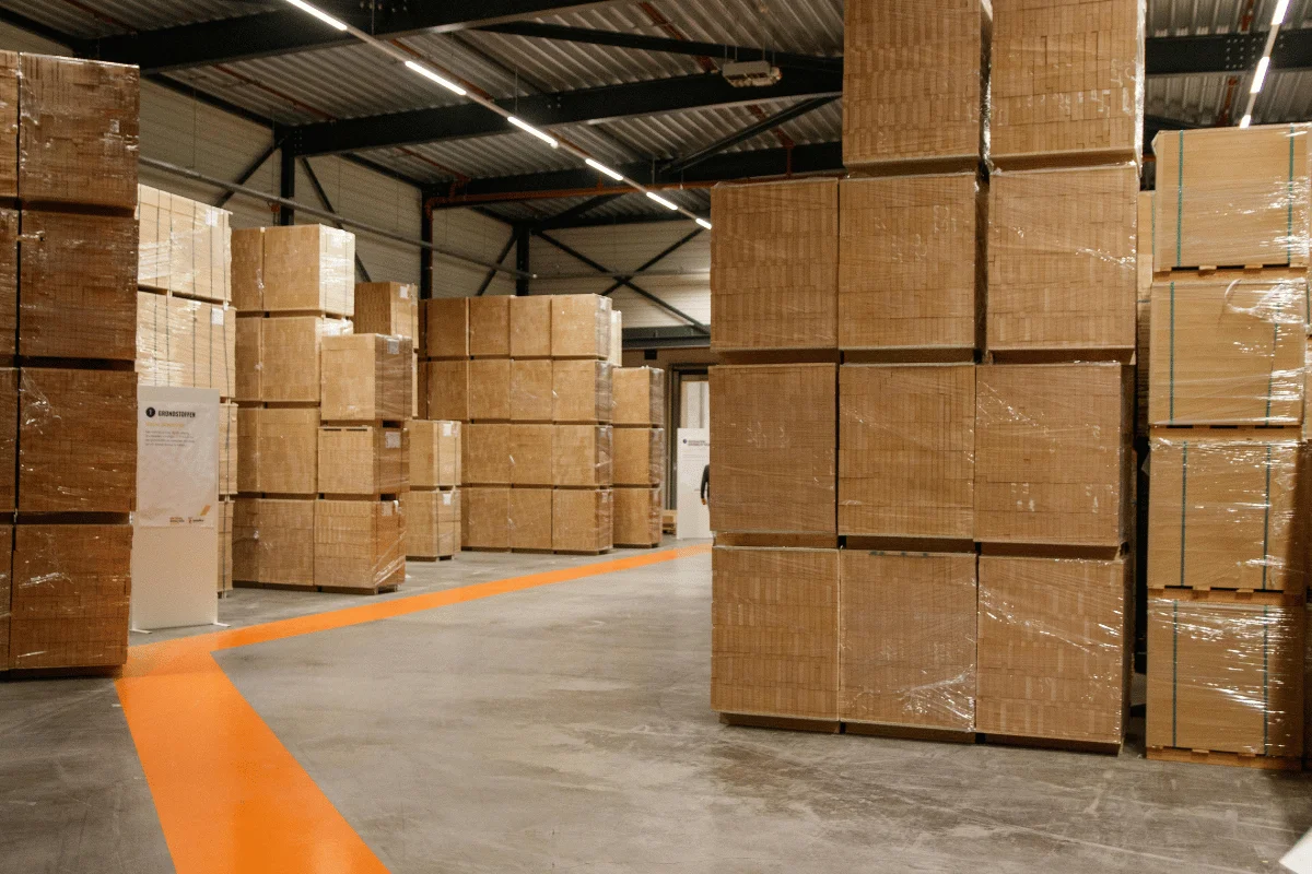 Spacious warehouse interior with stacks of sealed cardboard boxes on pallets, marked pathways, and industrial lighting, conveying a sense of order.