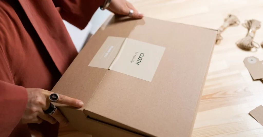 A person in a red garment holds a large, closed cardboard box labeled "CLOTH," on a light wooden table.