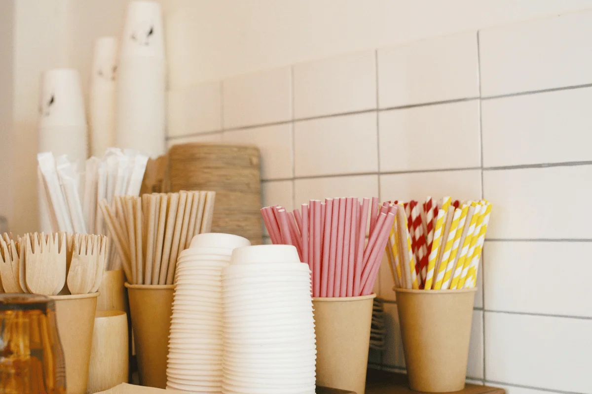 Disposable wooden utensils, stacks of white paper cups, and colorful paper straws in holders on a counter with a white tiled wall backdrop.