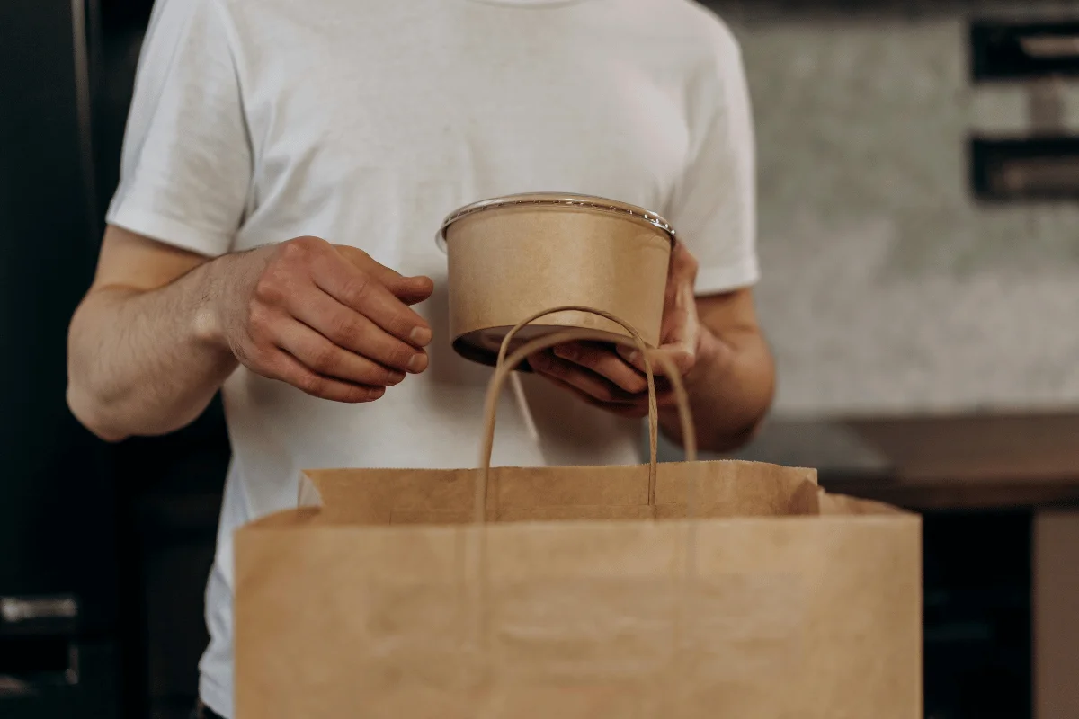 A person in a white t-shirt places a cardboard food container into a brown paper bag.