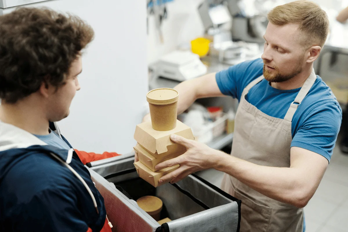 Man in apron hands food containers to a delivery person placing them in an insulated bag.