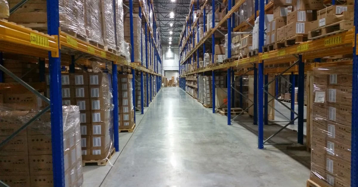 Industrial warehouse aisle with blue steel racks storing cardboard boxes and biopolymer types inventory under bright lights.