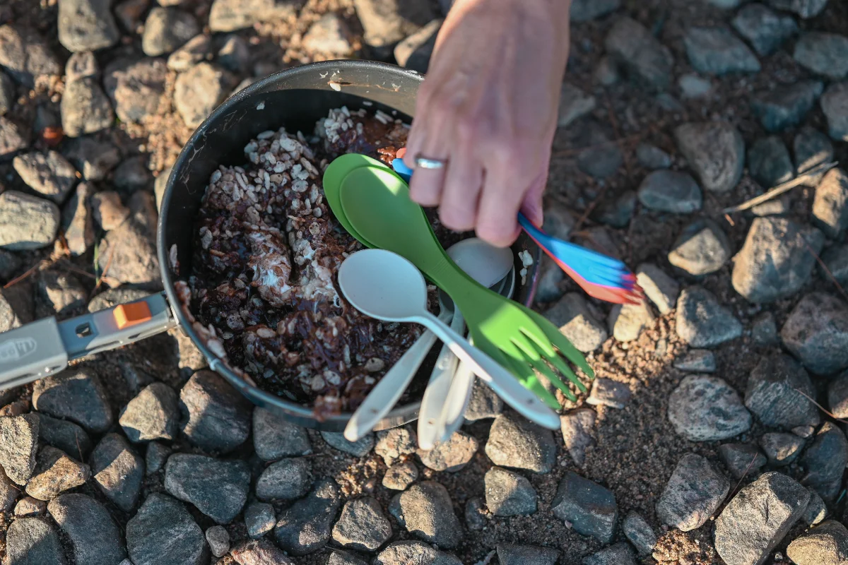 A hand holds a blue spork over a camping pot filled with a chocolatey rice mixture, with green and white plastic utensils resting inside the pan.