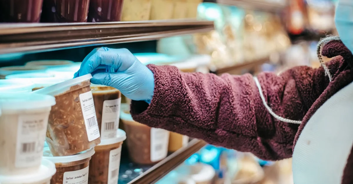 Hand in blue glove inspecting compostable packaging manufacturers' product labels on retail shelf display.