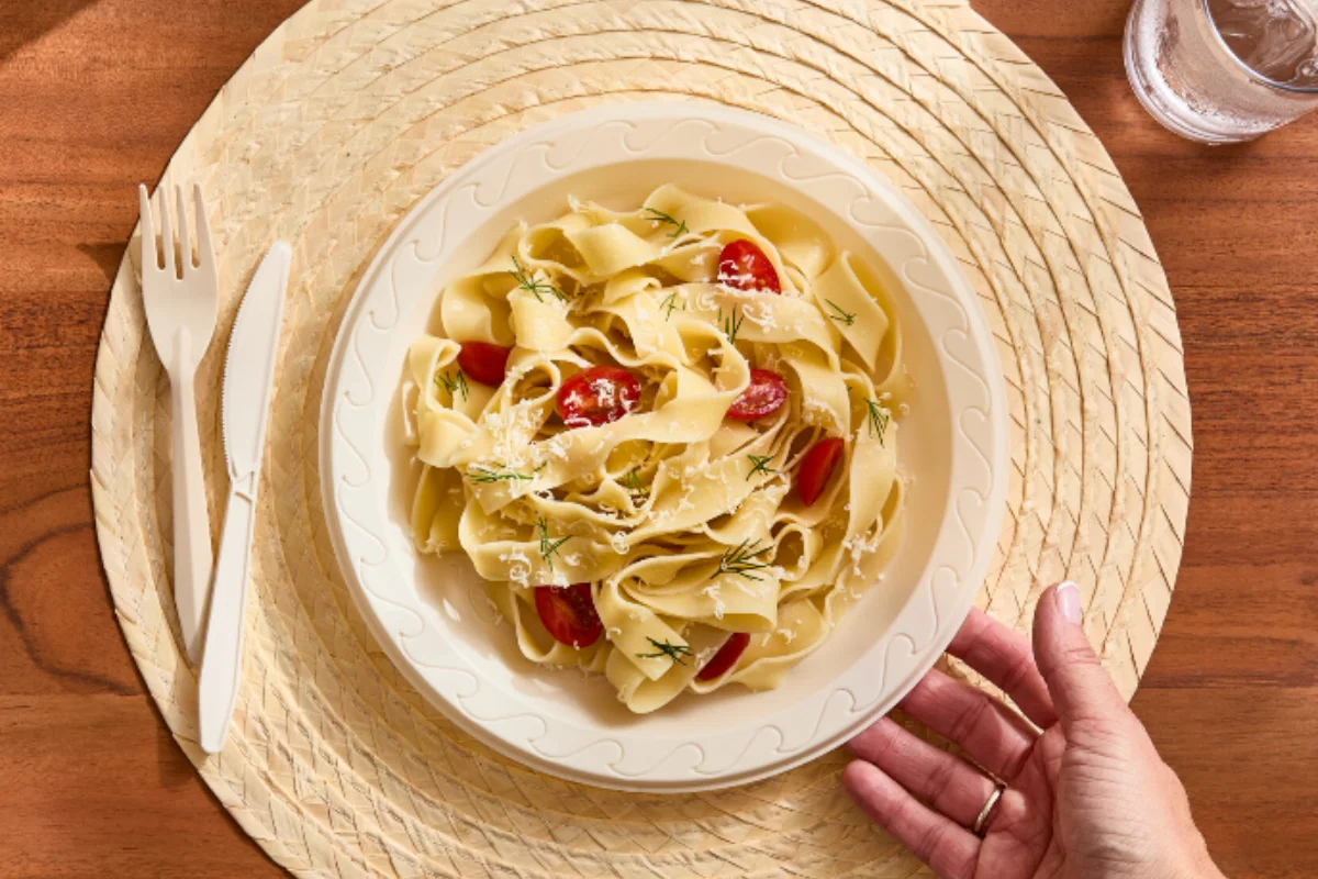 A plate of pappardelle pasta with cherry tomatoes, grated Parmesan, and herbs sits on a woven placemat. 