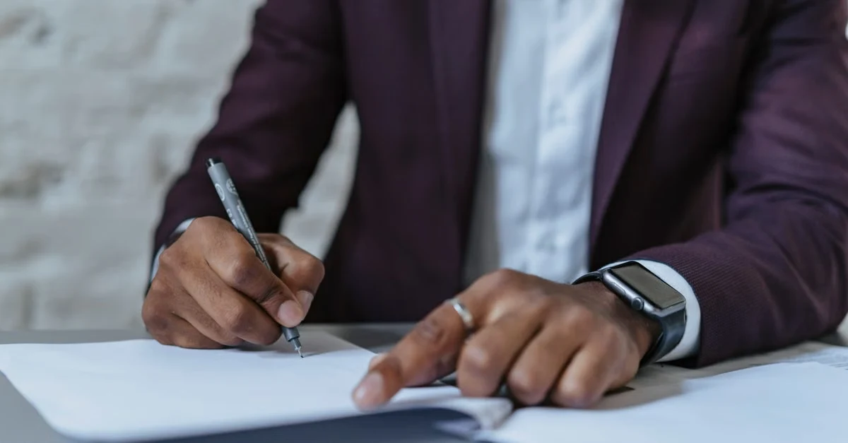 Professional in burgundy blazer signing contract document at desk with pen.