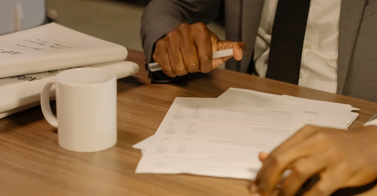 Business professionals reviewing compliance documents at wooden table with coffee mug and certification paperwork.