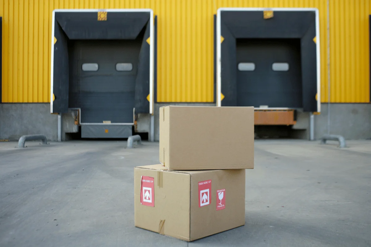 Two brown cardboard boxes with handling labels sit on the ground in front of a yellow warehouse with three closed loading dock doors.