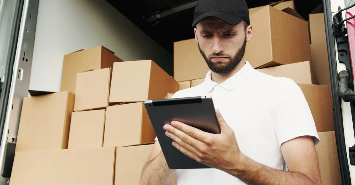 Delivery driver using tablet amidst stacked compostable packaging boxes assessing multi-state supplier capabilities.