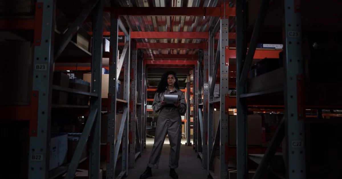 Worker standing in narrow aisle between industrial shelving units stocked with compostable products sourcing inventory.