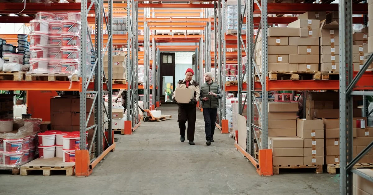 Workers walking through warehouse aisles with orange pallet racks storing compostable product inventory and stock boxes.