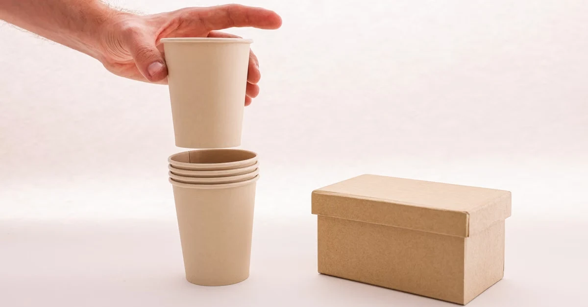 Hand stacking kraft paper cups showing compostable product shelf life and durability with cardboard box nearby.