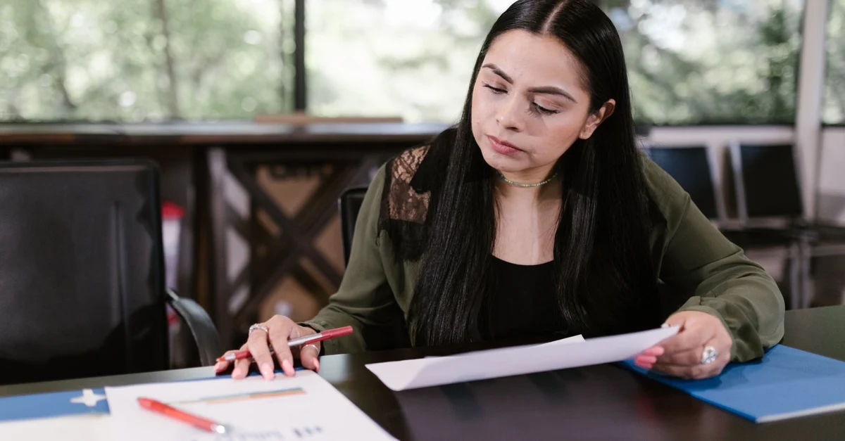 A woman with long black hair in an office setting is focused on reading a document.