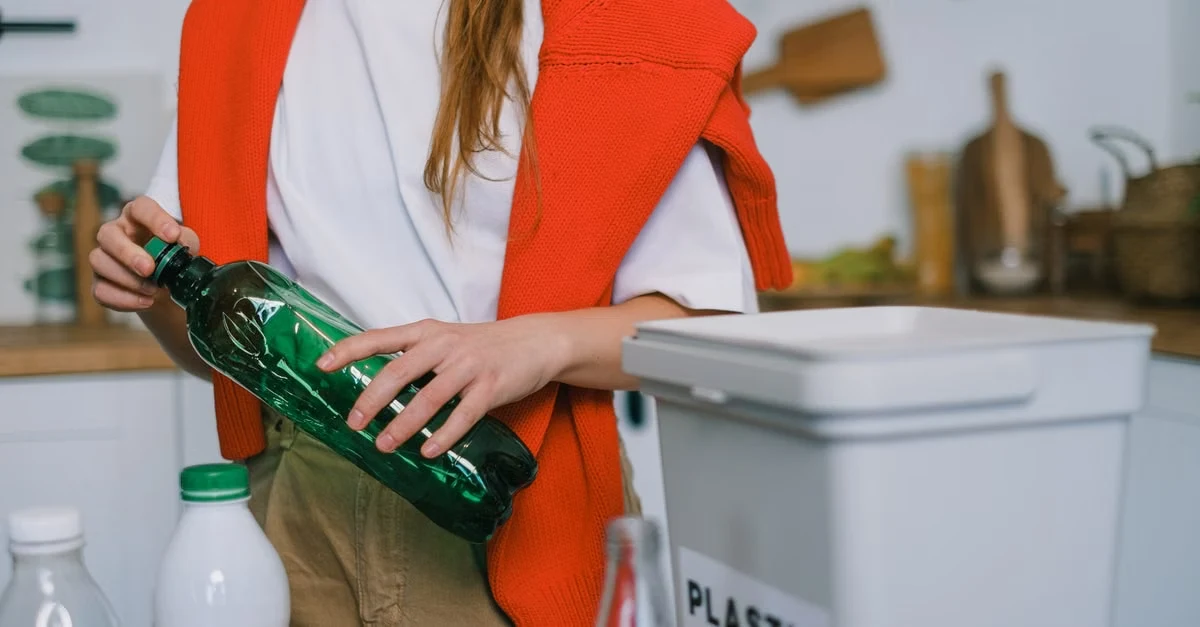 Woman placing green glass bottle into plastic recycling bin in kitchen, representing California SB 54 foodservice waste.