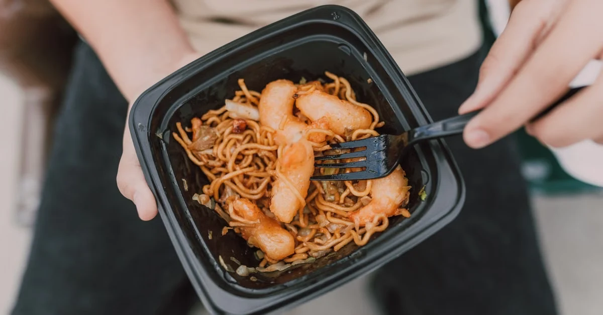 Person holding black biopolymer container with noodles and shrimp, demonstrating sustainable food packaging material.