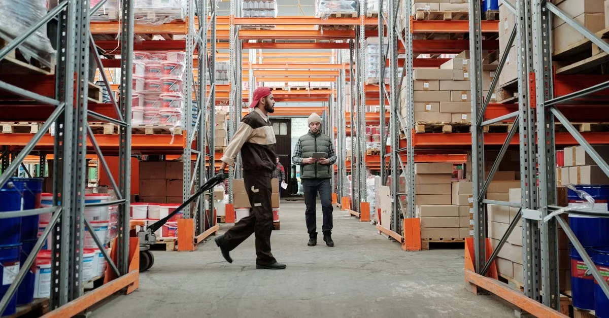 Workers checking inventory between tall metal shelving units with orange racks in warehouse distribution center.