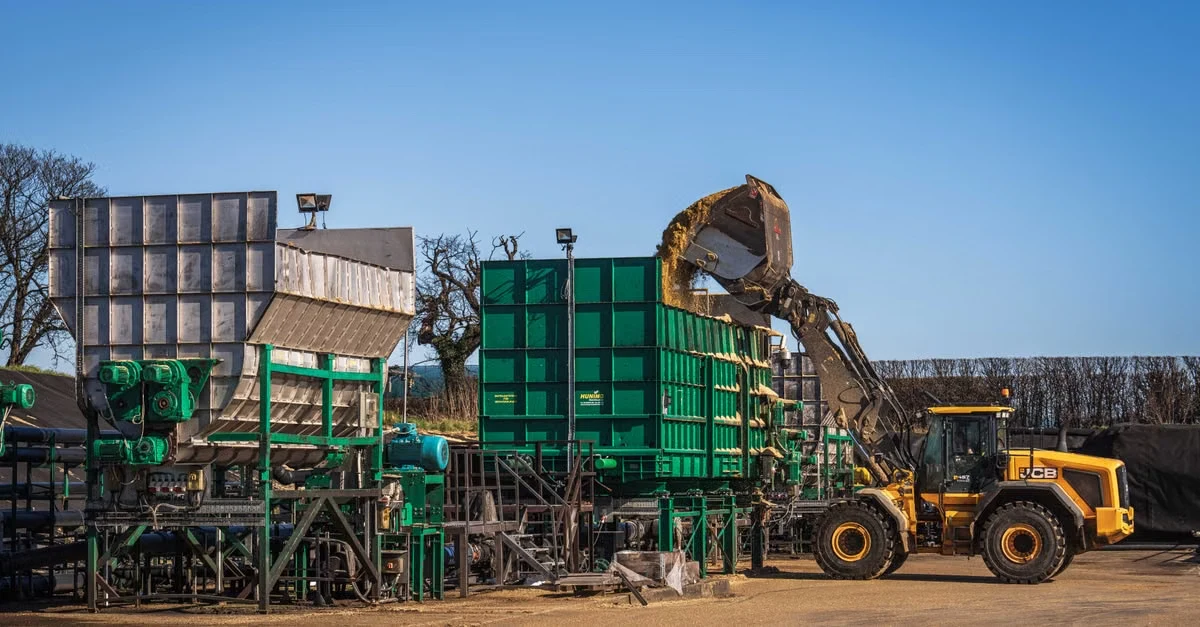 Commercial composting facility with green equipment, loader truck dumping organic waste into collection bins under clear sky.