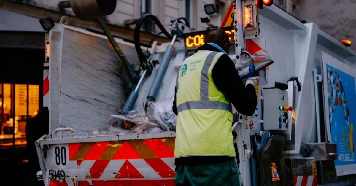 Worker in safety vest operating compostable waste collection truck with illuminated hazard lights at night.