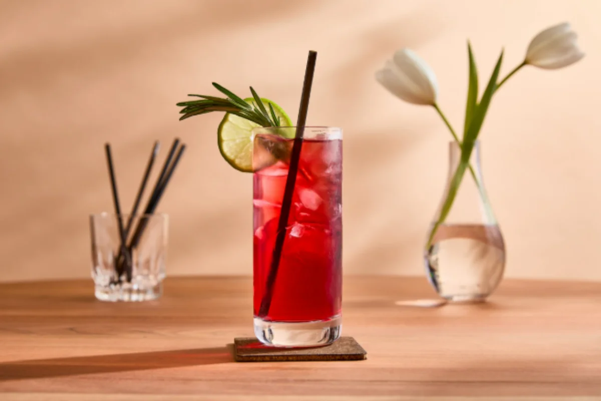 A tall glass of red iced cocktail with a lime slice, rosemary sprig, and dark straw on a coaster, with a vase of white tulips in the background.