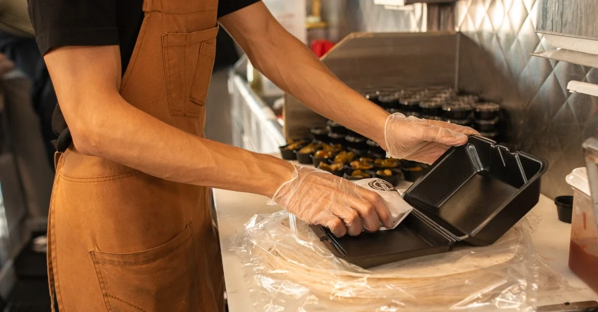 Worker in brown apron handling compostable material selection trays for greasy food service safely.