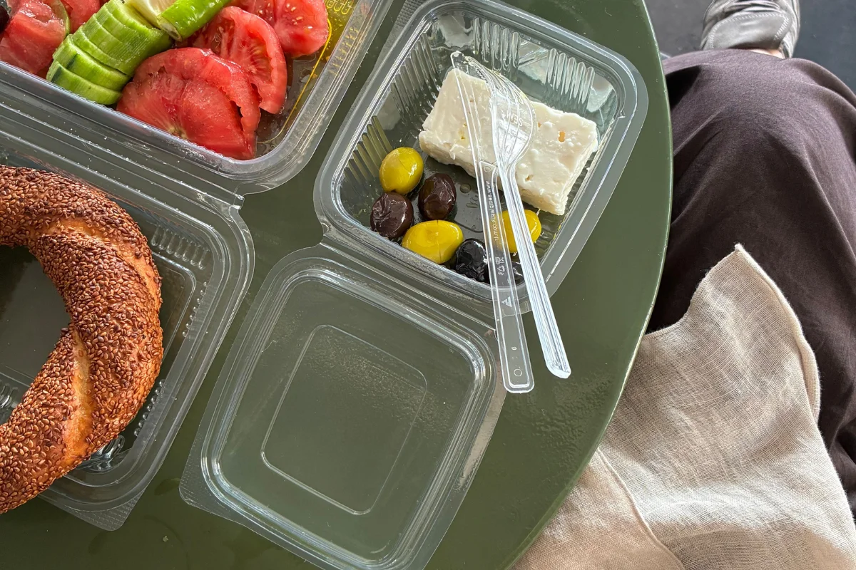 A sesame-covered ring bread, sliced tomatoes, cucumbers, feta cheese, and assorted olives in clear plastic containers on a round green table surface.