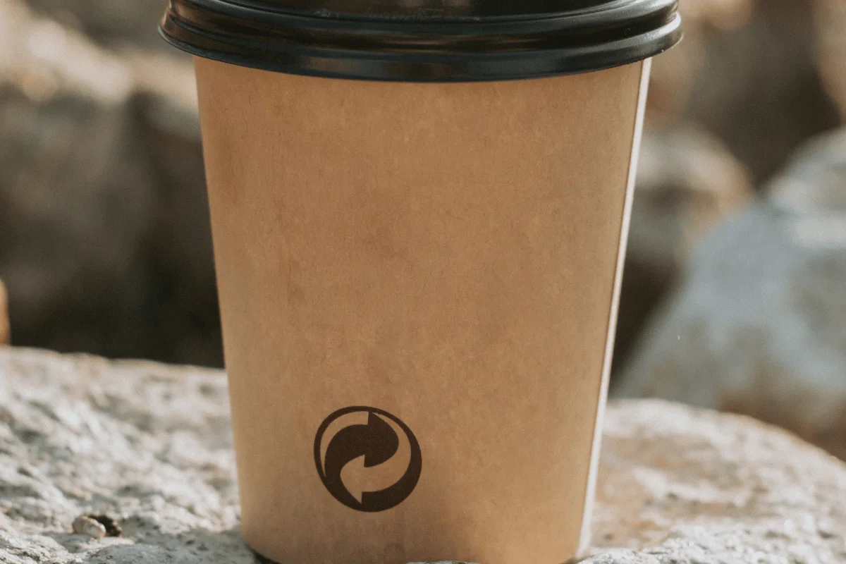 Brown disposable coffee cup with a black lid and circular logo rests on a sunlit rock outdoors, conveying a warm, natural mood.