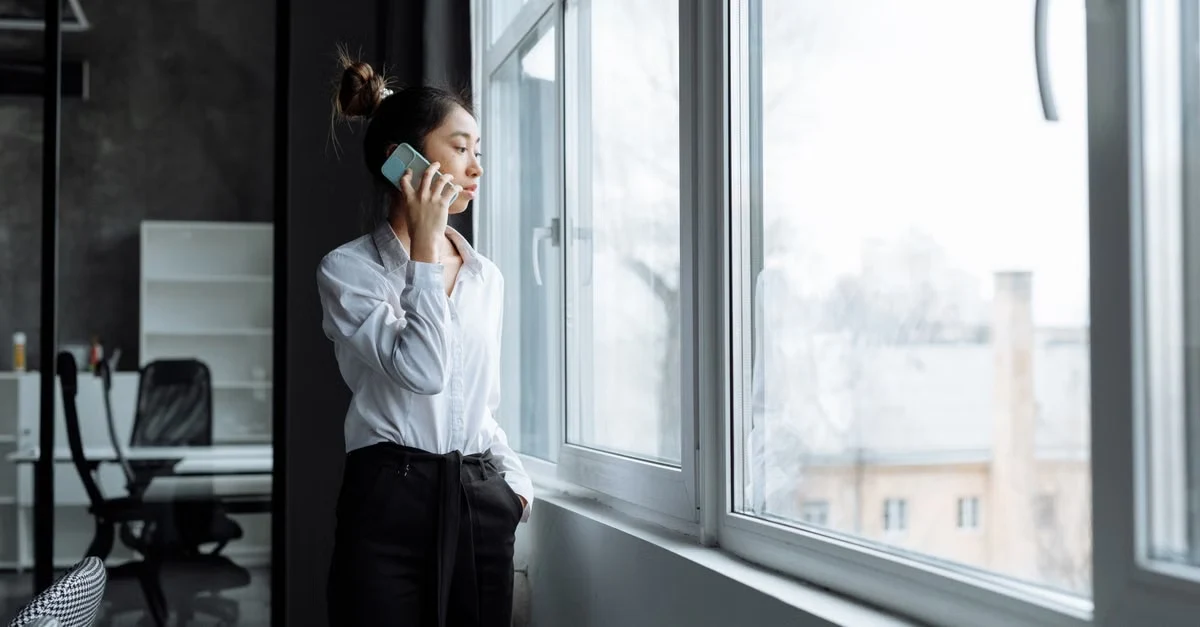 Professional woman with headphones calls from office window overlooking city, verifying compostable straws municipal.