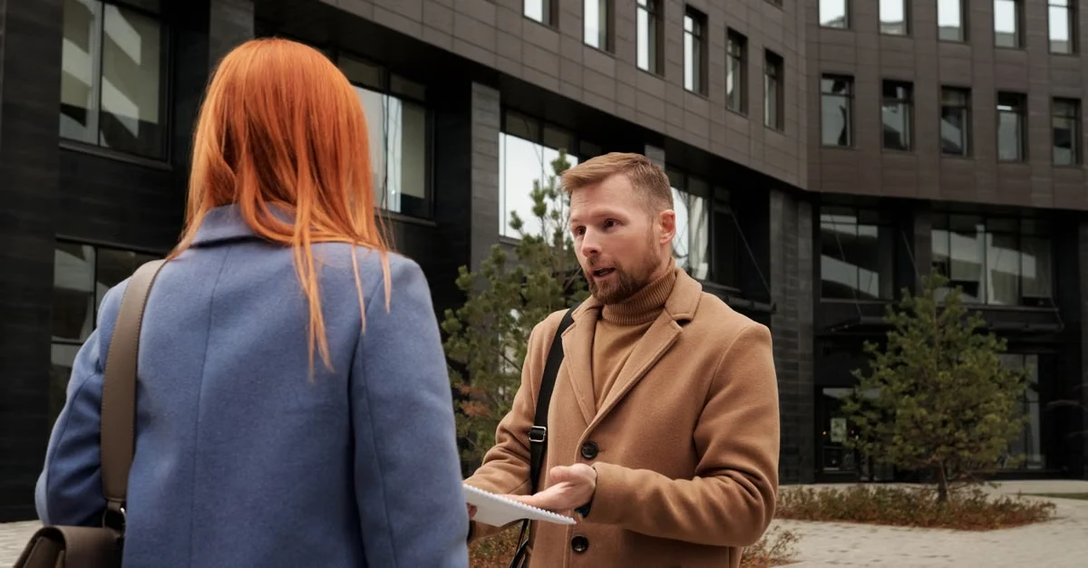 Man in brown jacket handing compostable straws information to woman near municipal building downtown.